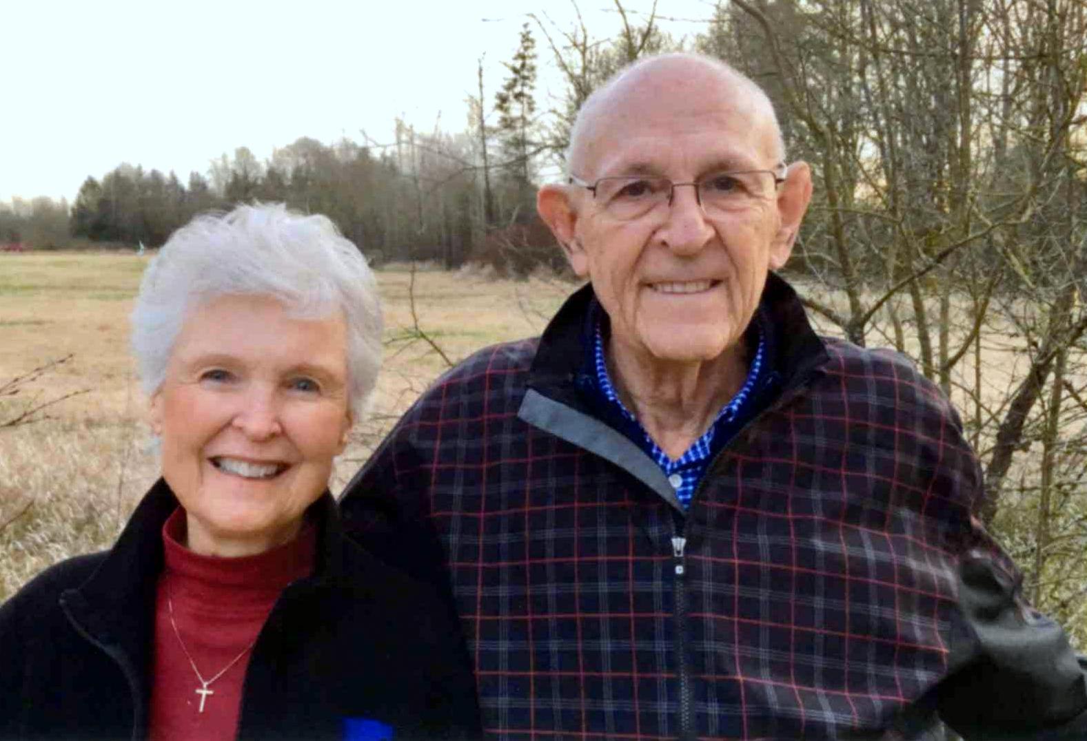 An elderly woman and man are standing outdoors in front of a field and trees. The woman has short white hair and is wearing a black jacket. The man, with glasses and short white hair, is sporting a plaid jacket. They appear to warmly welcome you to the peaceful surroundings of Presbyterian Manor.