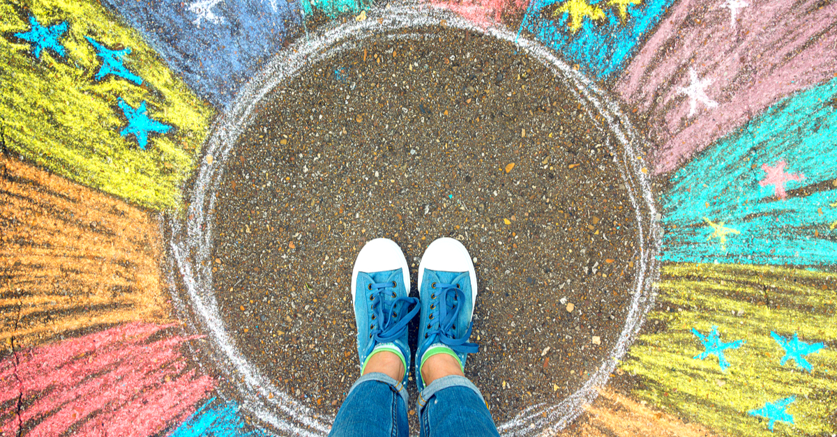 A person wearing blue sneakers stands on pavement adorned with colorful chalk drawings of stars and lines radiating outward from the center, symbolizing new beginnings.