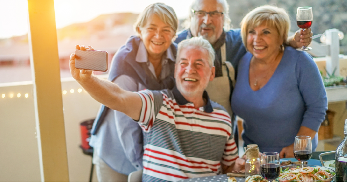 Four older friends smile and pose for a selfie at an outdoor dinner, where happiness fills the air with wine and food on the table.