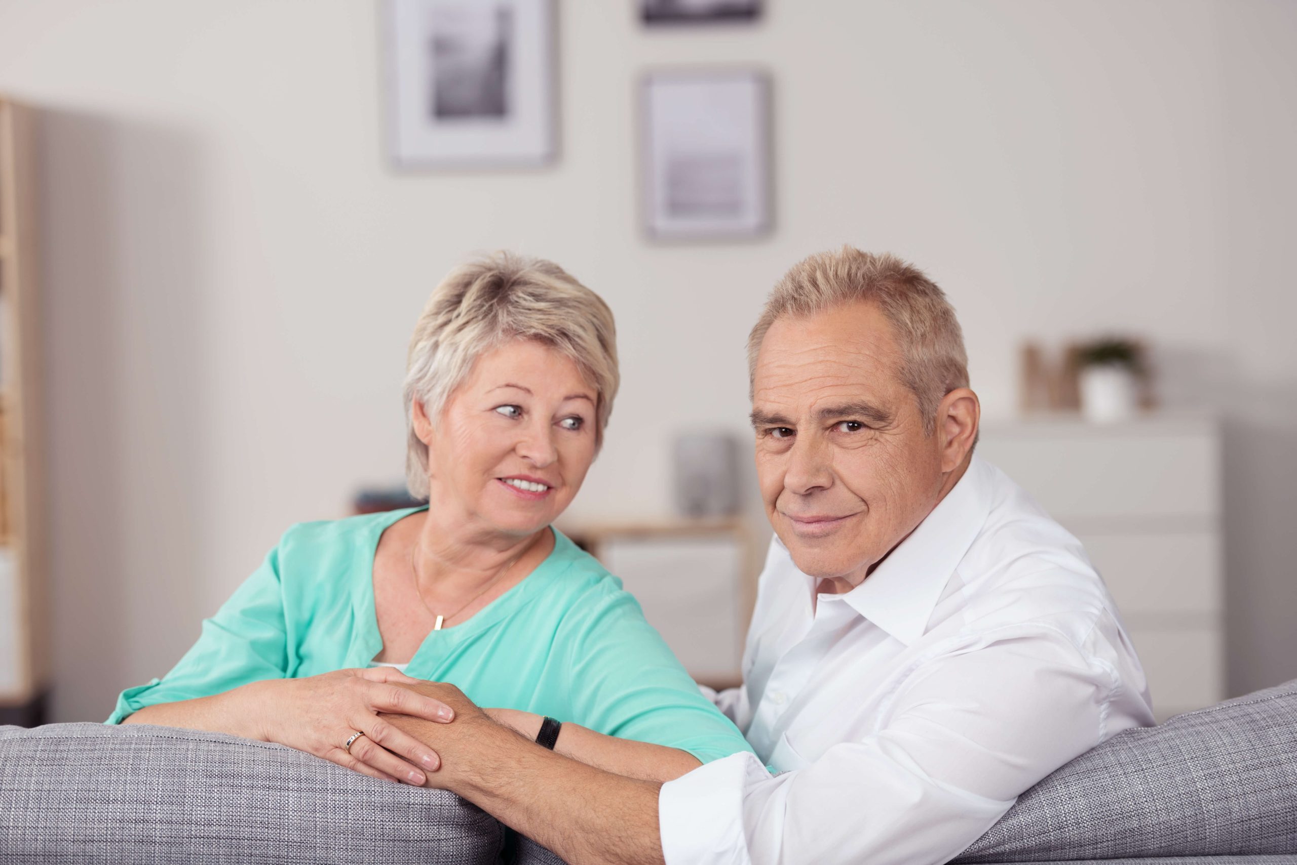 An older couple sits together on a gray sofa in a well-lit living room. The woman, wearing a turquoise top, smiles while the man in a white shirt looks at the camera. Framed pictures adorn the wall behind them, capturing moments that remind them it's always the right time to cherish senior living.