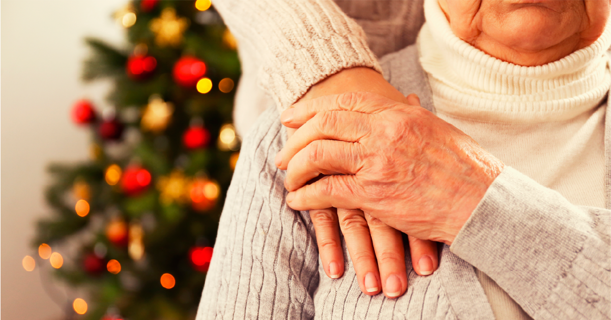 Two caregivers with interlinked hands stand in front of a decorated Christmas tree. One person gently rests their hand on the shoulder of the other, capturing the warmth and connection of the holidays.