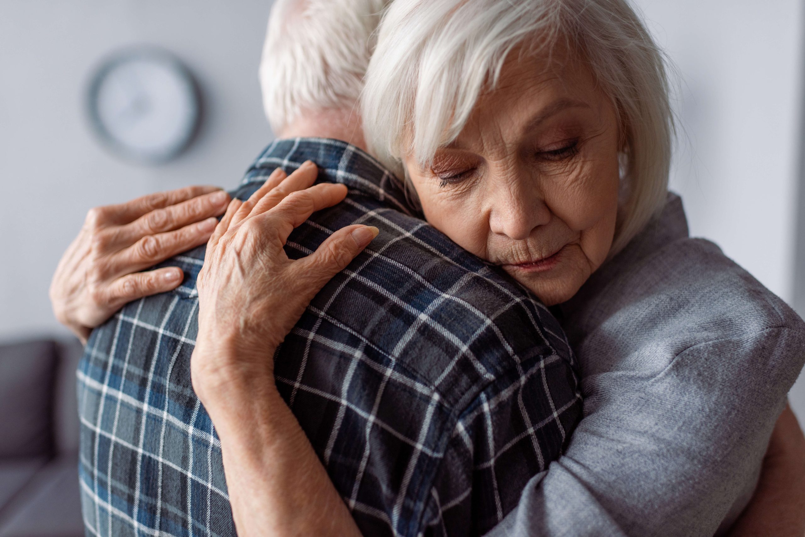 An elderly woman with gray hair embraces her partner, an elderly man with white hair wearing a plaid shirt. Both have their eyes closed, embodying the profound bond and caregiving that comes with facing the challenges of dementia together.