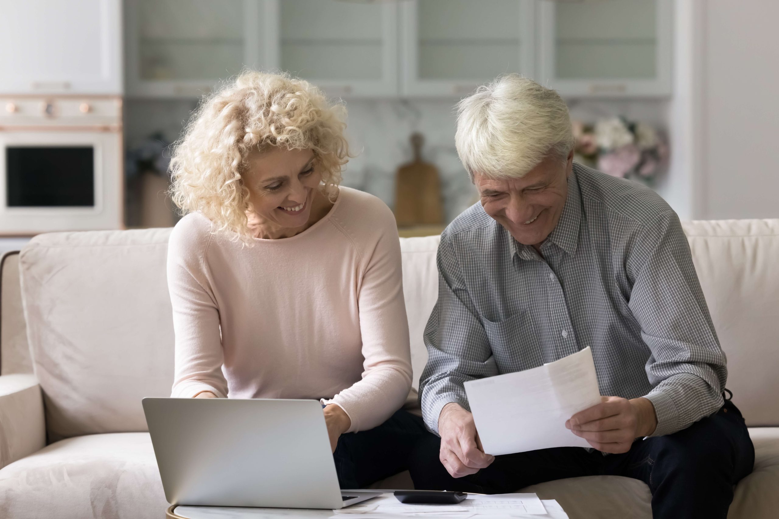 An older couple sits on a couch in their senior living community; the woman uses a laptop while the man looks at documents, both smiling.