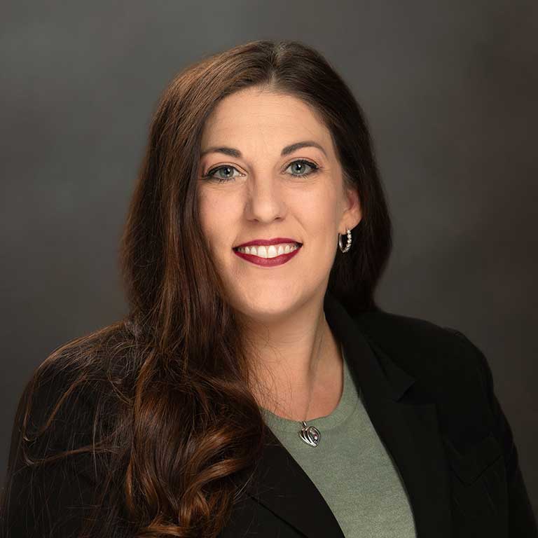 A woman with long brown hair and a dark blazer, smiling against a dark, blurred background.