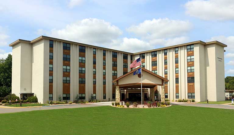 A multi-story building with numerous windows, a main entrance canopy, and two flags at the entrance, set against a backdrop of a grassy lawn and partly cloudy sky.