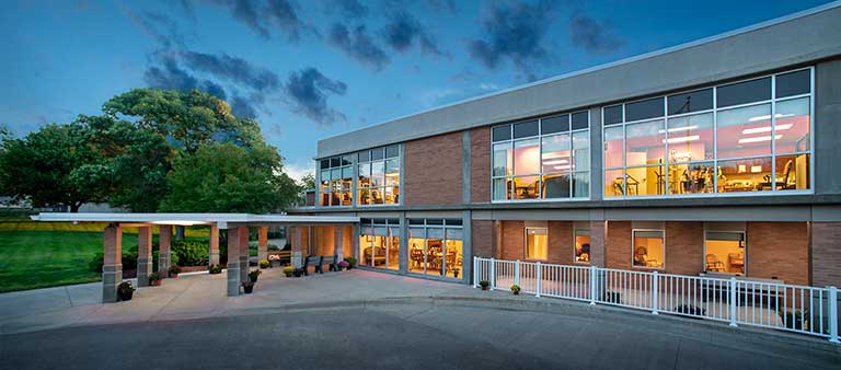 A two-story building with large windows is shown at dawn or dusk. The first floor exterior includes a covered entrance with pillars, and the interior is brightly lit, revealing various rooms.