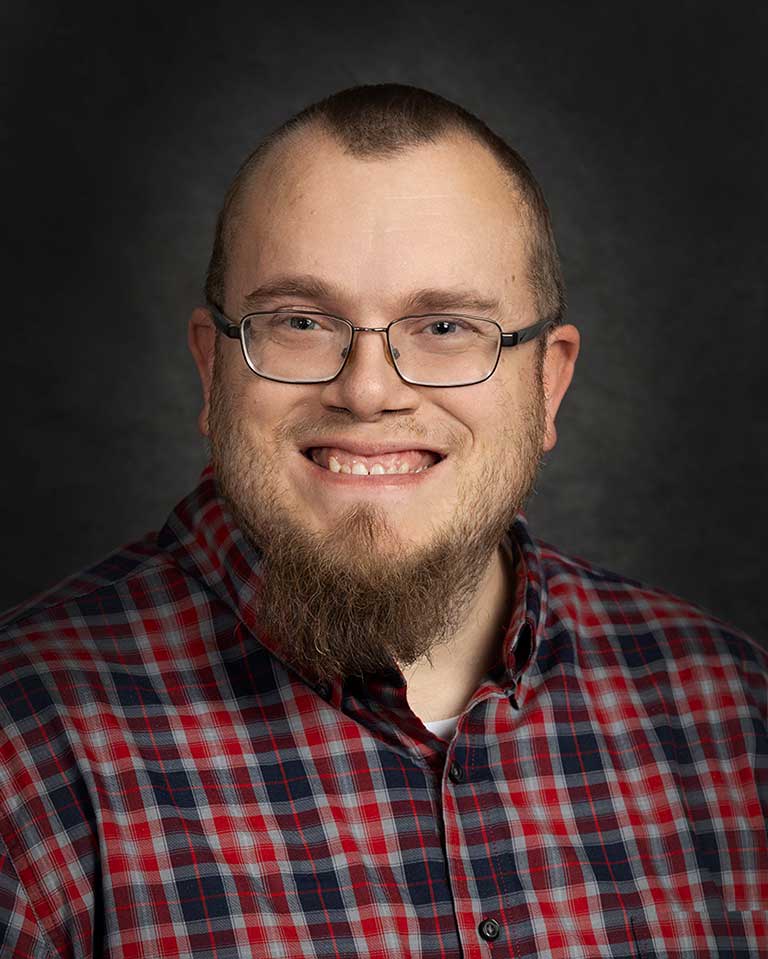 A person with glasses, a trimmed beard, and a shaved head is smiling. They are wearing a red and blue plaid shirt.