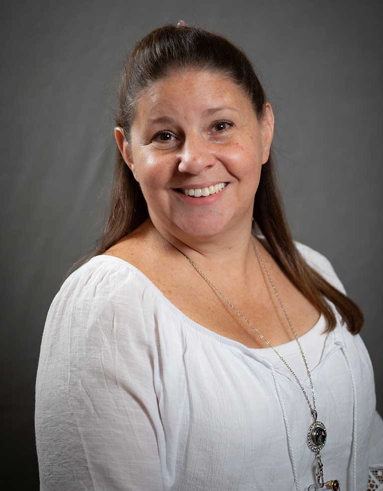 A woman with long brown hair is smiling at the camera. She is wearing a white blouse and a silver pendant necklace. The background is plain and gray.