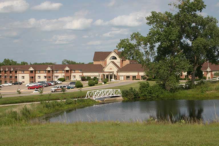 A large building with a brown roof, cars parked in front, a small bridge over a pond in the foreground, and trees surrounding the area, under a partly cloudy sky.