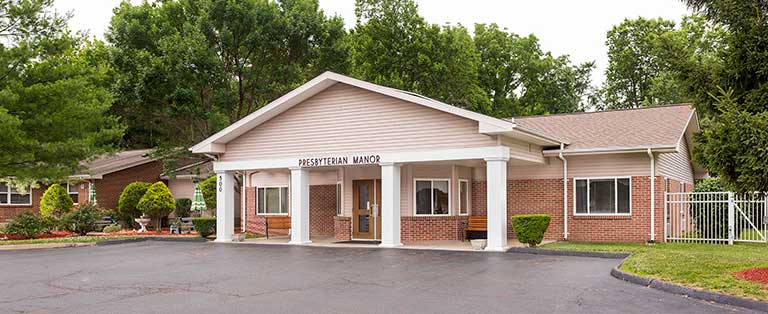 Single-story brick building with a beige, gabled roof and a sign reading "Presbyterian Manor" above its entrance. The building is surrounded by trees and has a parking lot in the foreground.