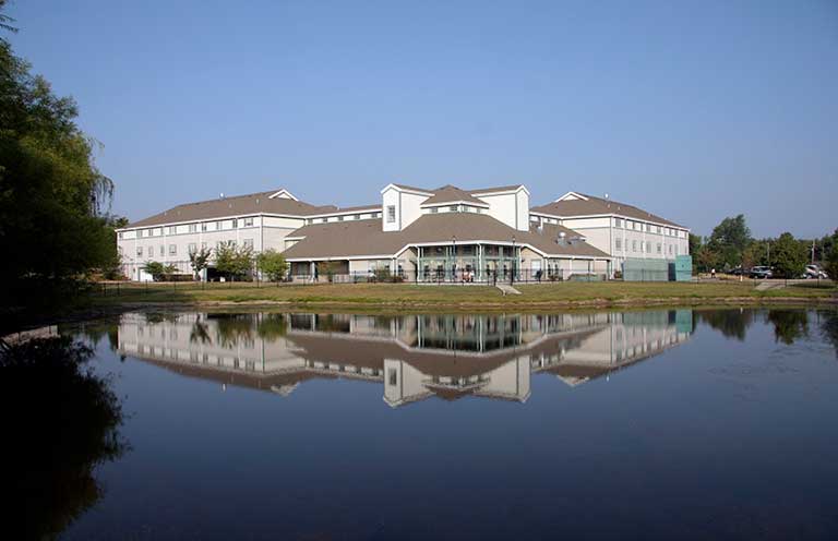 A large white building with multi-levels and a peaked roof is reflected in a calm body of water in the foreground, surrounded by greenery under a clear blue sky.