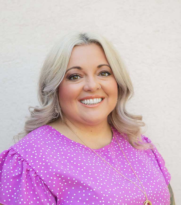 A smiling woman with blonde hair wearing a purple polka dot blouse and gold necklace stands in front of a plain light-colored background.