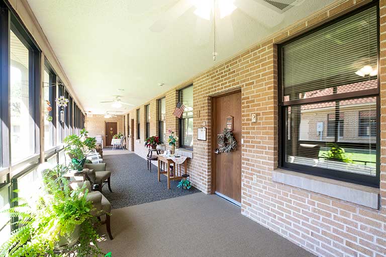 A brightly lit, enclosed hallway with brick walls, doors leading to individual rooms, and windows. The hallway features chairs, tables, plants, and decor.