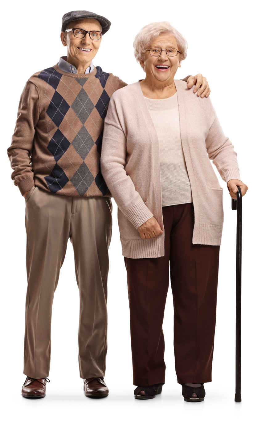 An elderly man and woman stand together, both smiling. The man wears a hat and argyle sweater, with one arm around the woman, who uses a cane and wears a light pink cardigan. They look content as they enjoy their time at the Colorado assisted living community.