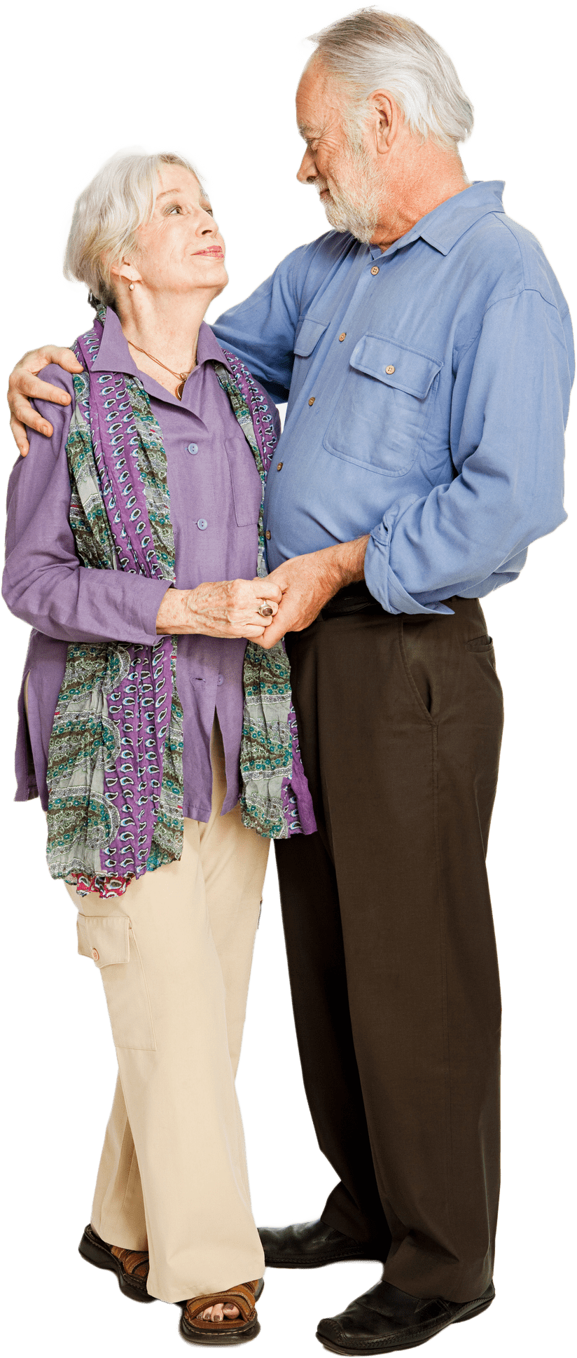 An elderly couple in senior living in Colorado looks at each other lovingly while standing in an embrace, with the man wearing a blue shirt and brown pants and the woman in a purple blouse and beige pants.