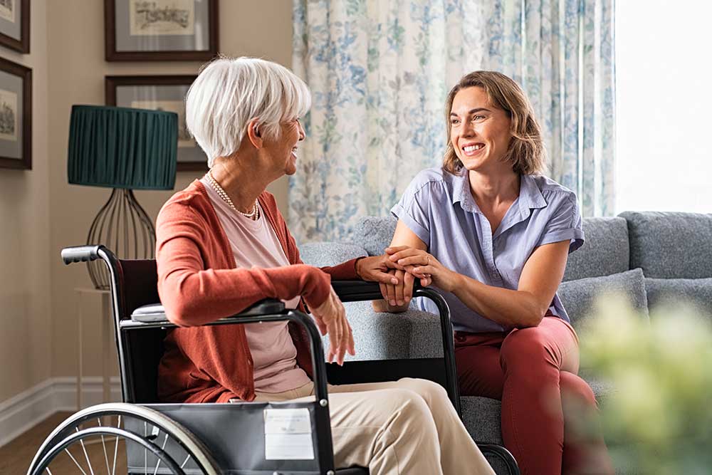 A woman in a wheelchair and a younger woman sit indoors, smiling and holding hands, suggesting a warm, caring interaction in a senior living facility in Kansas.