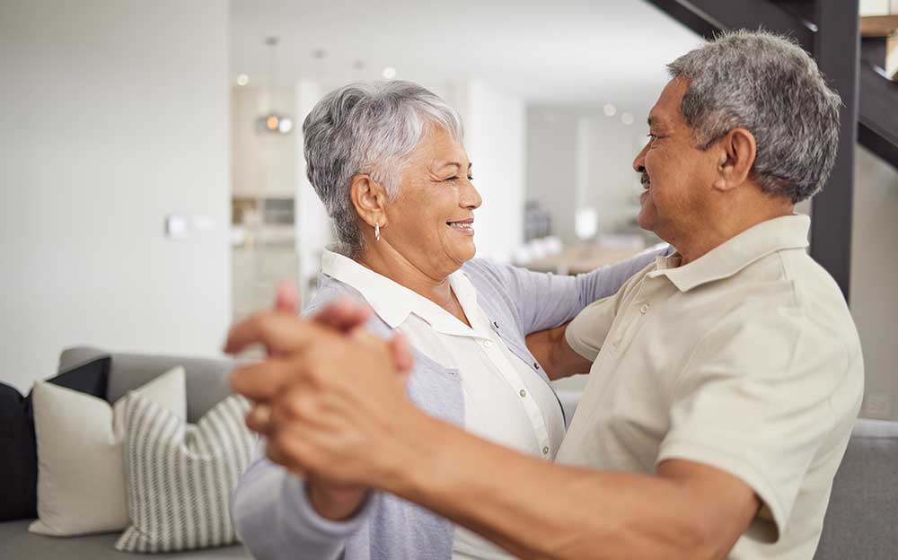 An older couple dances together in a bright living room, smiling and holding hands, enjoying their senior living in Colorado.