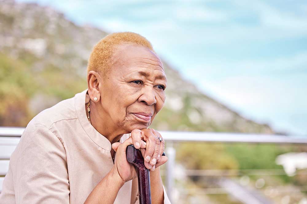 Elderly woman with short hair, wearing a light shirt, rests her chin on a cane while looking thoughtfully into the distance. The background features an outdoor setting with blurred natural scenery, evoking the serene environment of senior living in Missouri.