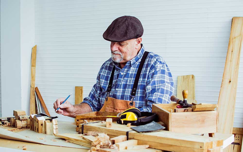 An older man wearing a flat cap and plaid shirt is working in a woodworking shop, surrounded by various tools and wooden pieces. This senior living in Kansas brings years of craftsmanship to life with each careful movement.