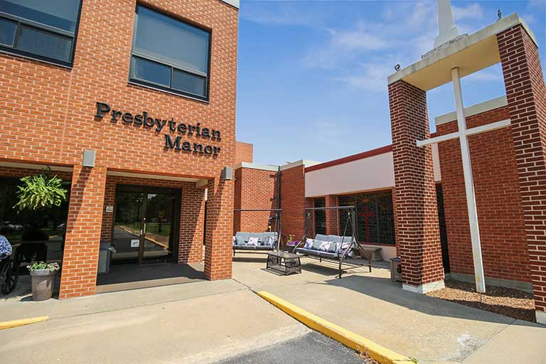 The image shows the exterior of "Presbyterian Manor," a brick building with large windows and a covered entrance area. A bench and some plants are visible near the entrance.