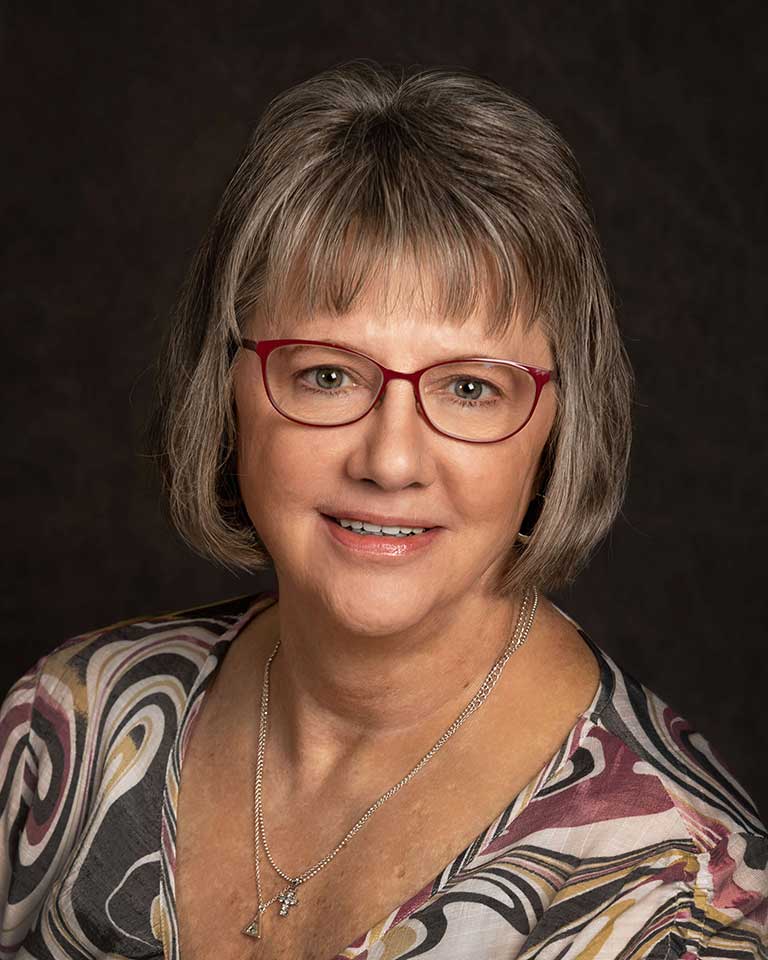 A woman with short gray hair and red glasses, wearing a patterned blouse and a necklace, smiles against a dark background.