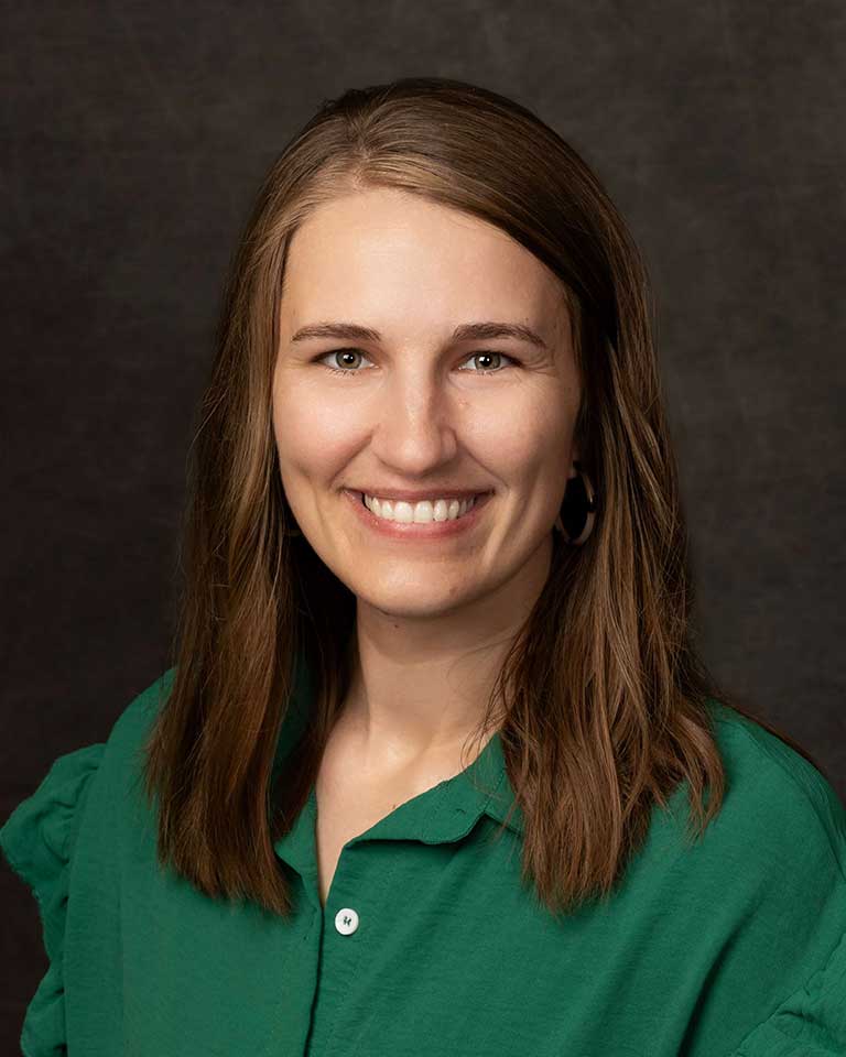 A woman with shoulder-length brown hair wearing a green shirt smiles against a dark background.