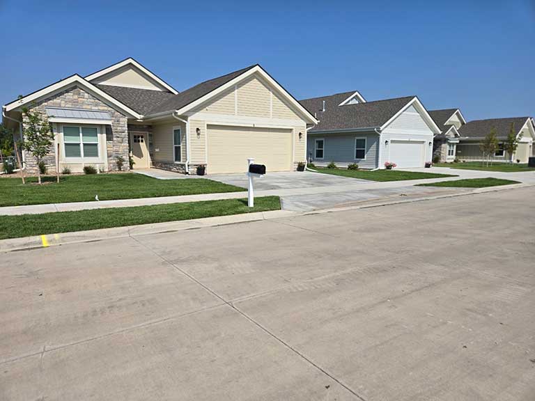 A row of newly built single-story houses with garages and driveways on a sunny day. The street is clean and there are no cars parked outside.