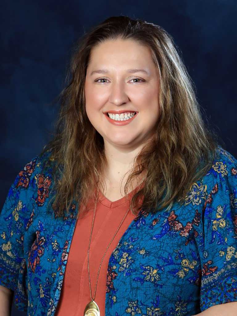 A woman with long brown hair smiles at the camera, wearing a blue floral-patterned cardigan over an orange top and a pendant necklace. The background is dark blue.