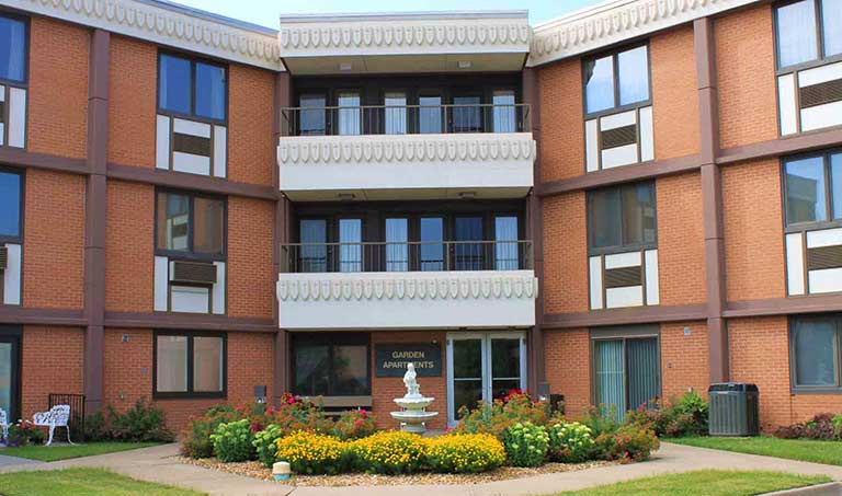 A three-story brick apartment building with a central entrance, labeled "Garden Apartments," surrounded by landscaped gardens and a white fountain.