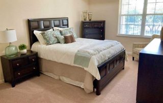 A bedroom with a wooden bed frame, two nightstands, a tall dresser, a patterned chair, and a window with blinds. The bed is made with white bedding and decorative pillows.