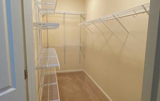 A clean, empty walk-in closet with beige carpet, beige walls, and wire shelf organizers on both sides under a ceiling light.