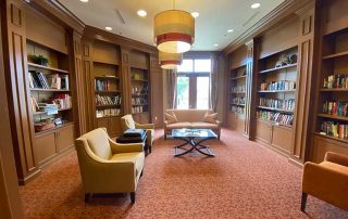 A well-lit library room with wooden shelves filled with books, comfortable seating, and a coffee table in the center. Large windows at the back let in ample natural light.