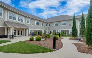 A two-story residential building with a courtyard featuring pathways, outdoor seating, plants, and trimmed bushes under a blue sky with scattered clouds.