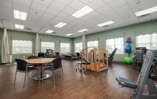 A spacious physical therapy room with exercise equipment, a wooden staircase apparatus, a round table with chairs, and large windows allowing natural light.