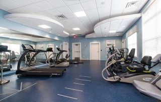 A clean gym with treadmills, recumbent bikes, and exercise equipment on a blue floor. Mirrors cover one wall, and windows line another, allowing natural light into the room.