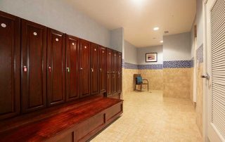 A locker room with wooden lockers and tiled walls. A picture and a chair are positioned at the far end of the room, which has beige tile flooring and recessed ceiling lights.