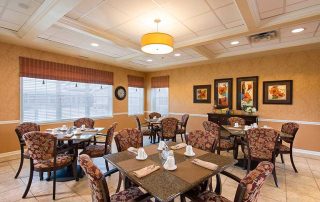 A dining room with several tables and chairs set with napkins and small white cups. The room has floral paintings on the walls and large windows with blinds. A ceiling light fixture hangs above.