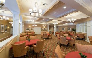 A dining area with red and beige upholstered chairs, red tablecloths, decorative lighting, artwork on the walls, and patterned carpets.