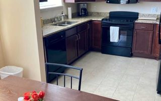 A kitchen with dark brown cabinets, a black stove, oven, and microwave, black dishwasher, sink, and countertops. A dining table holds a red flower arrangement in the foreground.