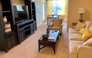 A living room features a beige couch, two armchairs, a wooden coffee table, a TV on a black entertainment center, and a dining area with a table and chairs near yellow curtains.