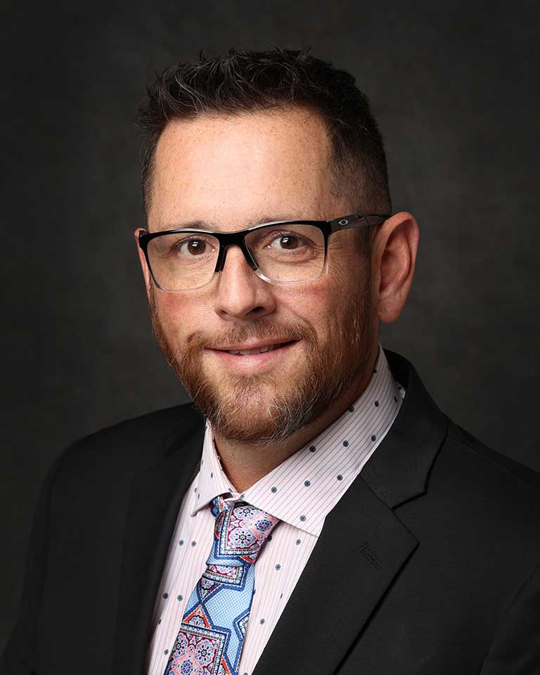 A man with short hair and glasses wearing a suit jacket, a white dotted shirt, and a patterned tie is posing for a professional headshot against a dark background.