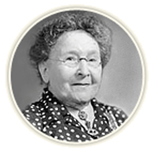 Black and white image of an elderly woman with curly hair and glasses, wearing a polka dot blouse, smiling slightly.