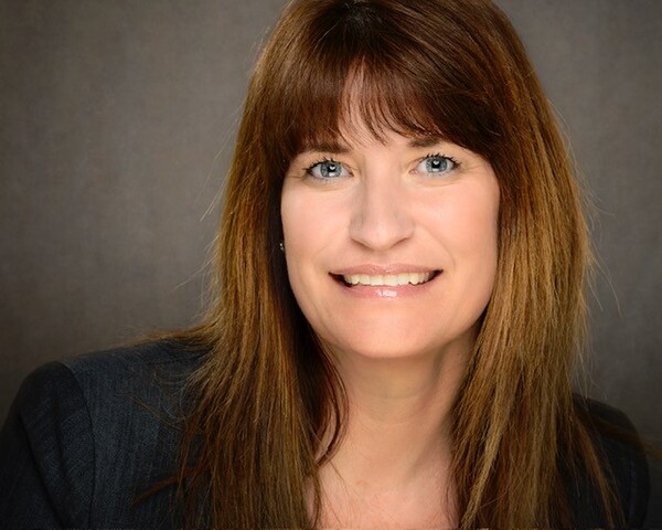 A woman with straight brown hair and blue eyes is smiling. She is wearing a dark jacket against a neutral background.