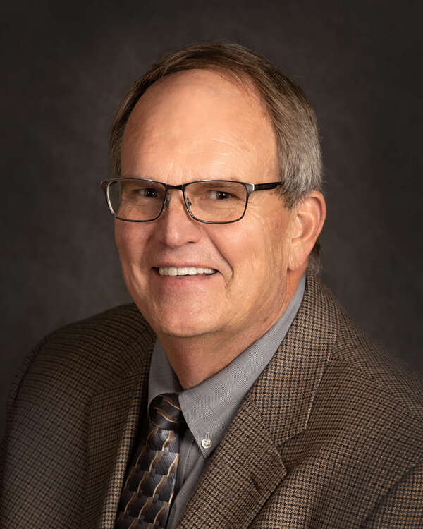 A man with short hair wearing glasses, a checkered brown blazer, a gray collared shirt, and a patterned tie smiles against a dark background.