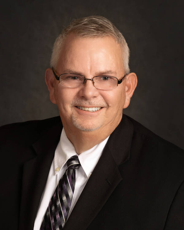 A smiling man with short gray hair and glasses is wearing a dark suit, white shirt, and striped tie. He poses against a dark background.