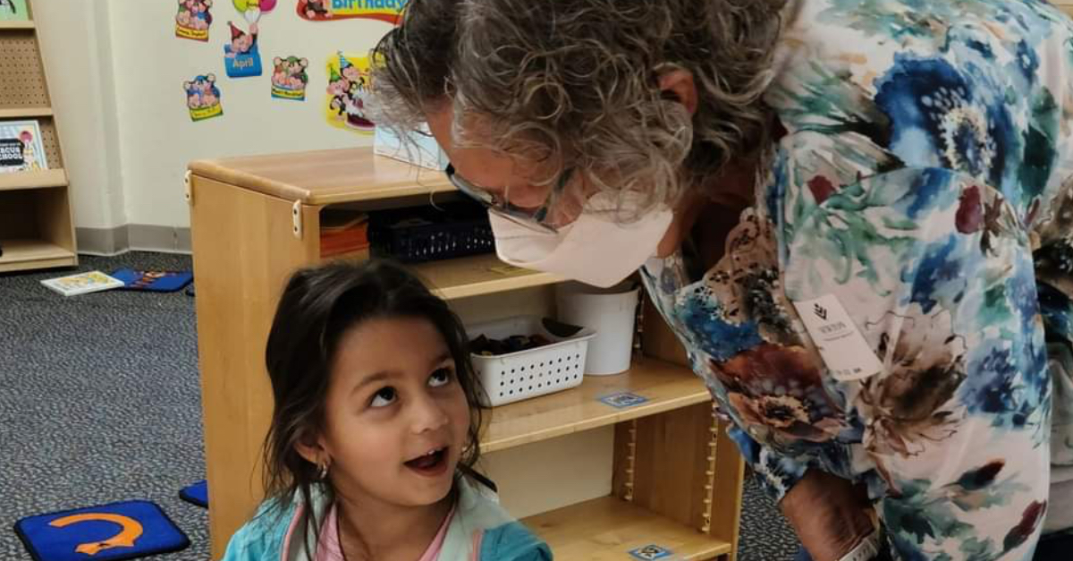 At Newton’s Apple-A-Day Preschool, an adult with curly gray hair wearing a mask and a floral shirt leans toward a young child in the classroom. The child, with long dark hair and a surprised expression, looks up at the adult during the anniversary celebration.