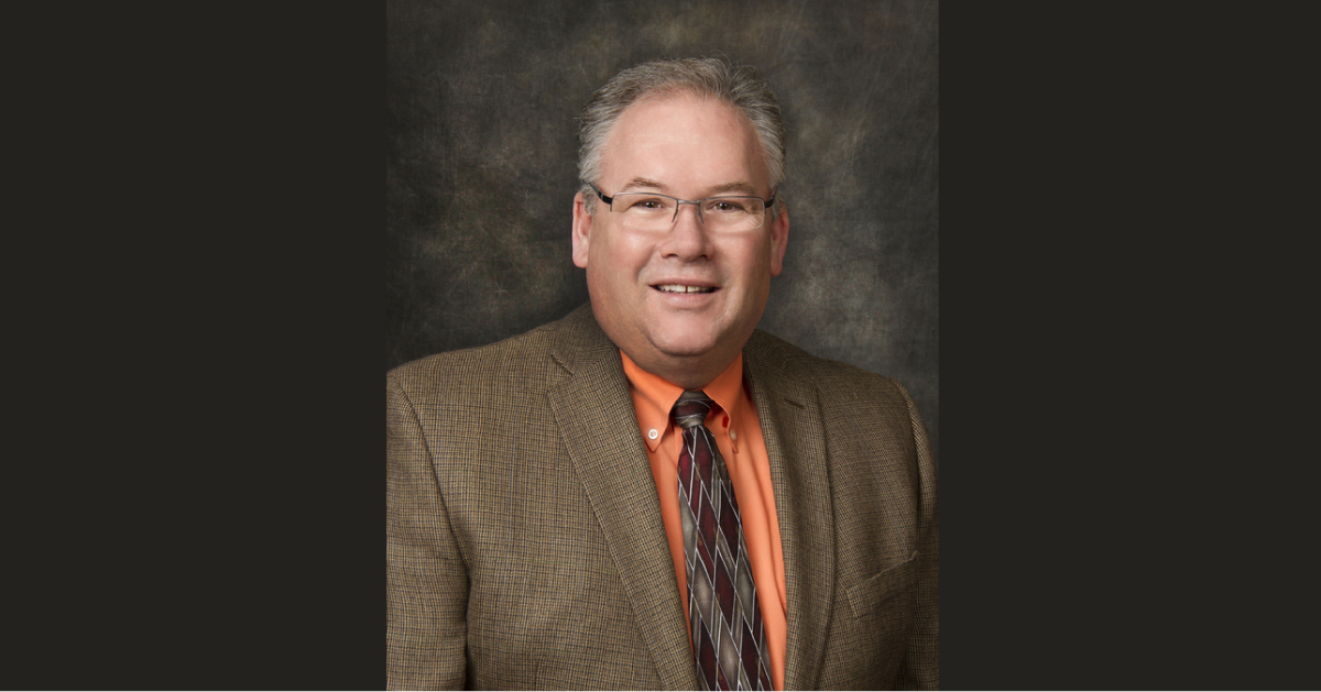 A man with gray hair and glasses wearing a brown blazer, orange shirt, and striped tie poses against a dark, textured background, exuding a sense of wisdom and calm reminiscent of a kind chaplain.