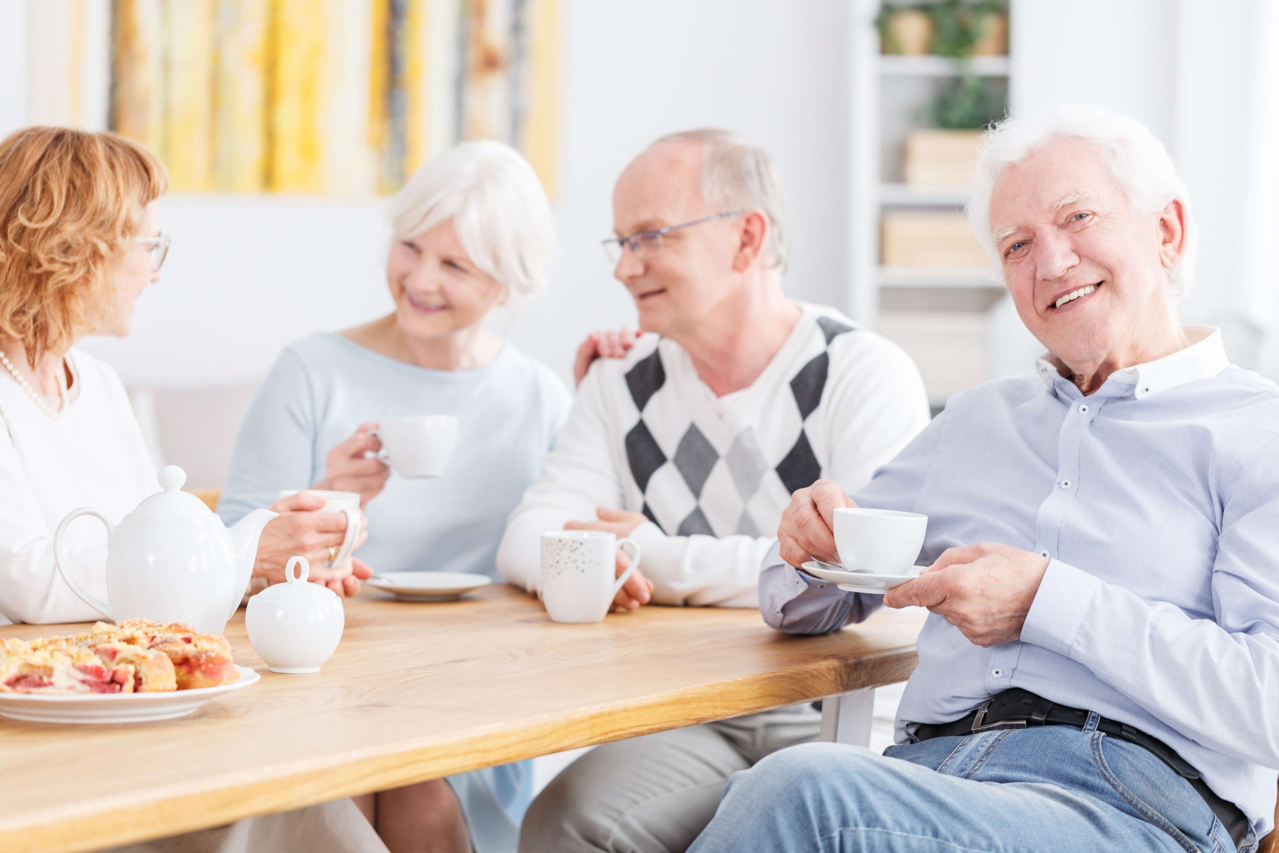 Four elderly adults are sitting around a wooden table, enjoying tea and pastries. They are engaged in conversation about independent living, with one person smiling directly at the camera.