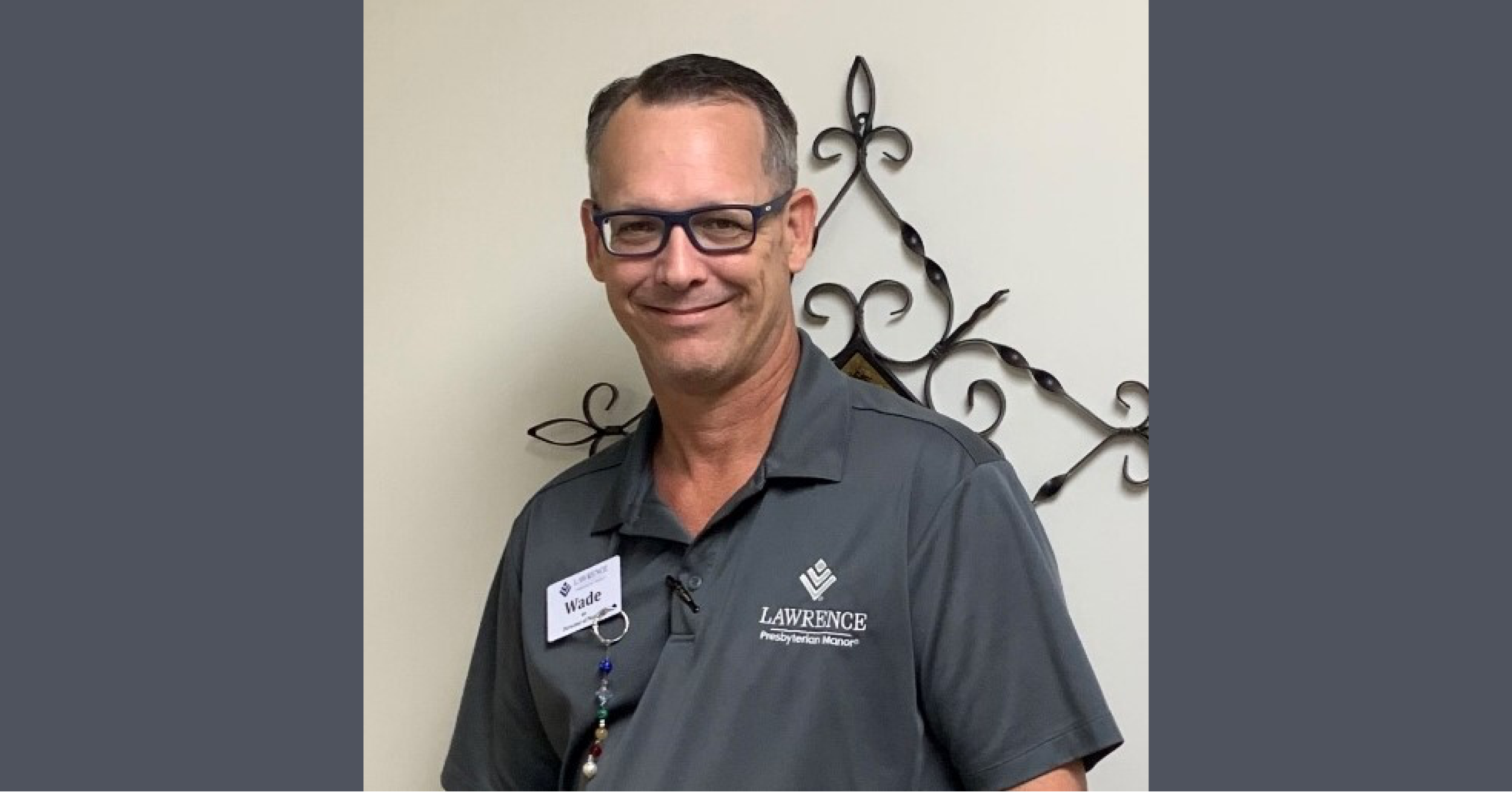 A man wearing glasses and a gray polo shirt with a name tag stands in front of a wall with decorative ironwork at Lawrence Presbyterian Manor, where life comes full circle.