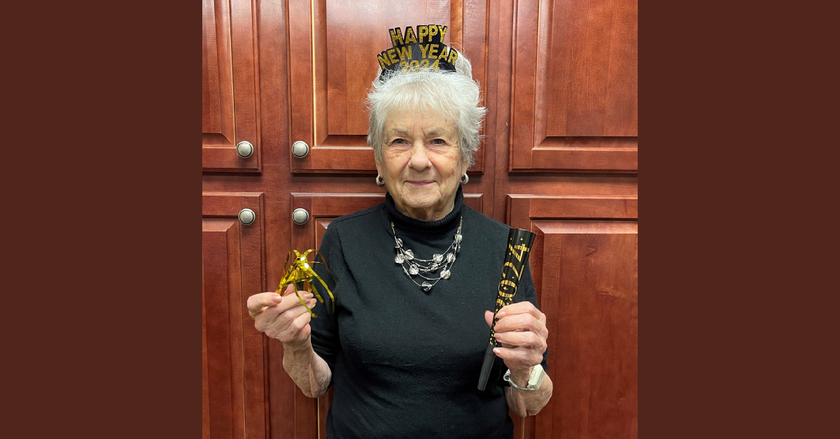 An elderly woman with white hair, Dorothy Pallister, wears a "Happy New Year" headband and holds a 2024 noise maker and a gold decoration in front of brown wooden cabinets. Know Your Neighbor and celebrate the festive spirit with her!